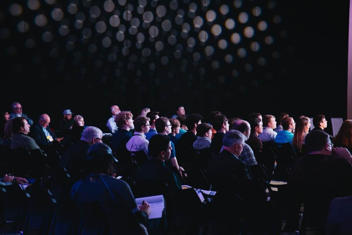 A seated conference audience faces a stage under a ceiling of small round lights