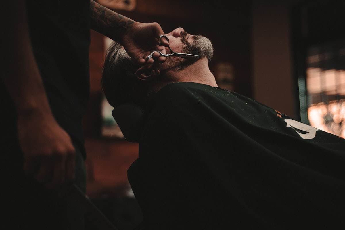 A barber trims a reclining client's beard with scissors in a dimly lit shop