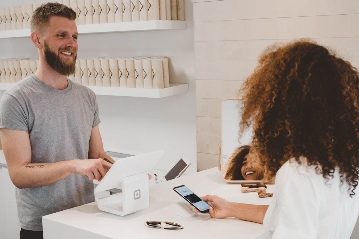 A shop assistant at a white counter taps a tablet register while a customer waits with her phone in hand